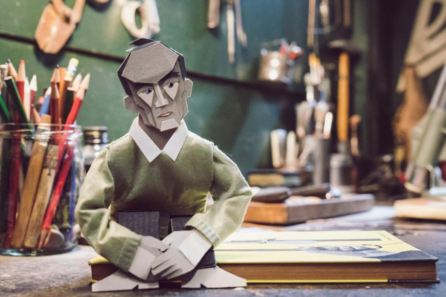Eine kleine Pappfigur sitzt auf einem Arbeitstisch mit Kunstmaterialien und vielen Stiften.