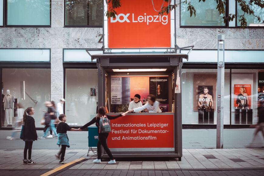 Zwei Personen in einer Info-Box in einer Fußgängerzone. Eine der Personen reicht einen Flyer an vorbeigehende Kinder.