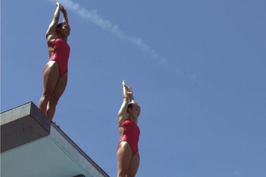 Zwei Frauen in roten Badeanzügen stehen bereit zum Absprung auf einem Sprungturm.