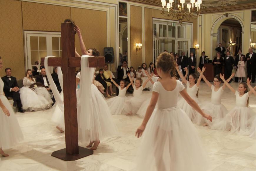 Mehr als ein Dutzend Mädchen tanzt in weißen Ballettkleidern vor einem großen Holzkreuz, das mit Schleiern dekoriert ist.