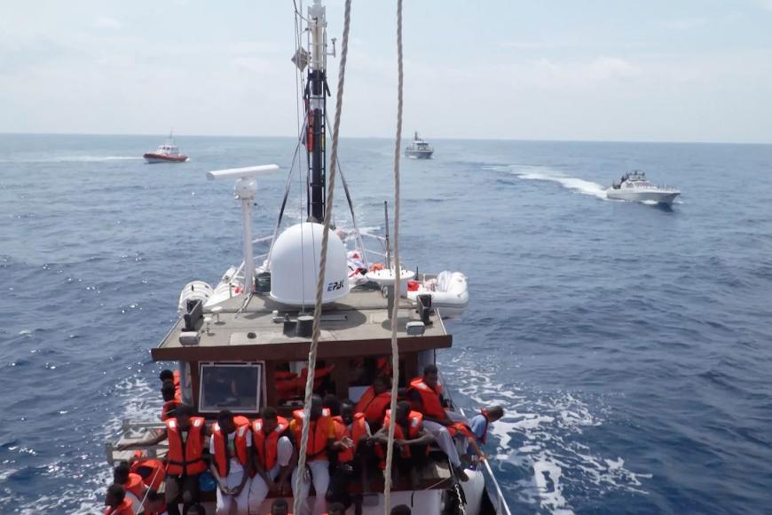 Ein Rettungsschiff mit vielen Personen in roten Rettungswesten an Deck im Meer. Im Hintergrund drei kleinere Boote.