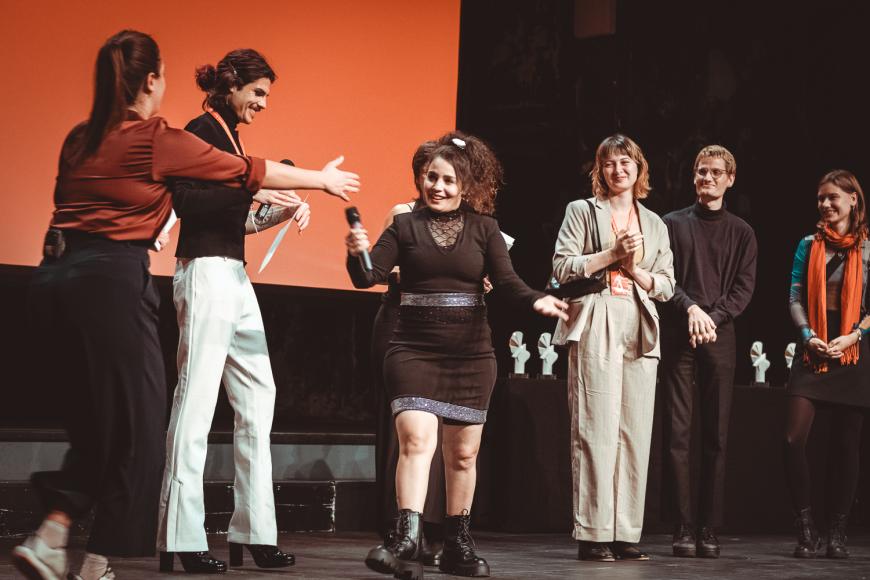 Sechs Personen auf einer Bühne vor einer roten Leinwand. Alle lächeln, einige applaudieren. Die Frau in der Mitte strahl und geht einen Schritt nach vorne. 
