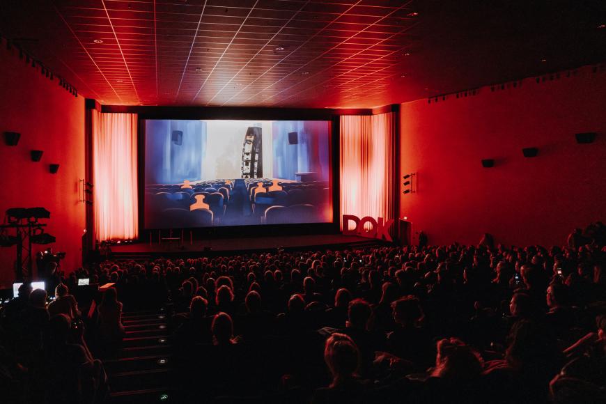 A fully occupied cinema hall, lit up in atmospheric red light. An excerpt from the festival trailer can be seen on the screen: small red weights, also sitting in a cinema.