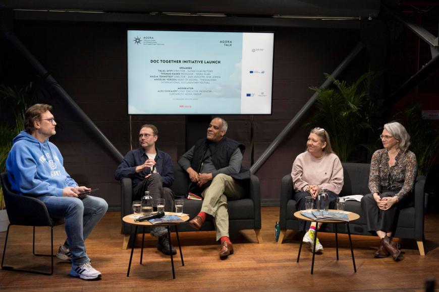 Vier Männer und eine Frau sitzen auf einer Bühne mit einer Leinwand im Hintergrund, die das Thema „Doc Together Initiative Launch“ anzeigt.