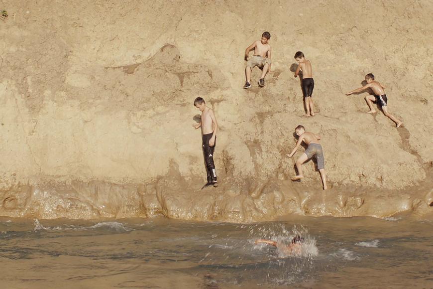 Four young boys in bathing clothes climb down a golden cliff towards water, one boy has just jumped in the water