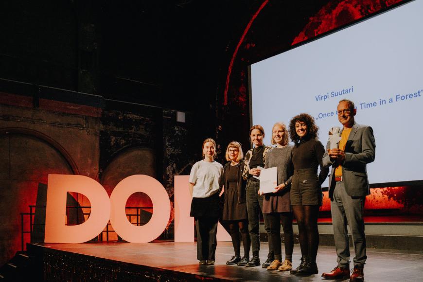 Die fünfköpfige Publikumsjury steht auf der Bühne bei der Preisverleihung, neben ihnen hält eine Person aus dem Gewinnerfilmteam grinsend die Goldene Taube Statue in den Händen