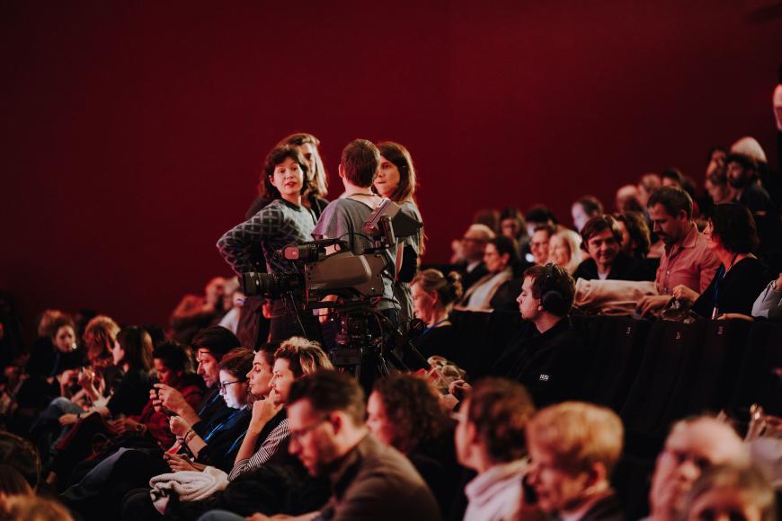 In einem vollen vollen Kinosaal stehen in einer Sitzreihe vier Personen im Gespräch. Eine Person dreht sich gerade zur Kamera und schaut freudig und überrascht