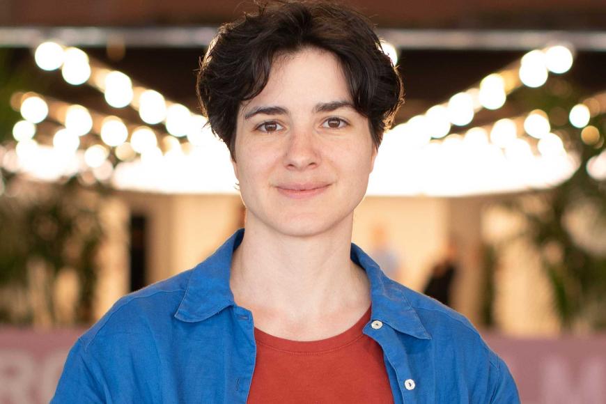 Portrait of Babette Dieu with short hair in a blue shirt and red t-shirt standing in front of blurry lights