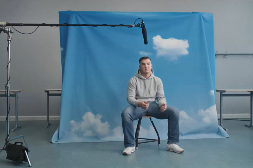 Jugendlicher sitzt vor aufgestelltem Wolken-Hintergrund. Über ihm hängt ein Mikrofon.