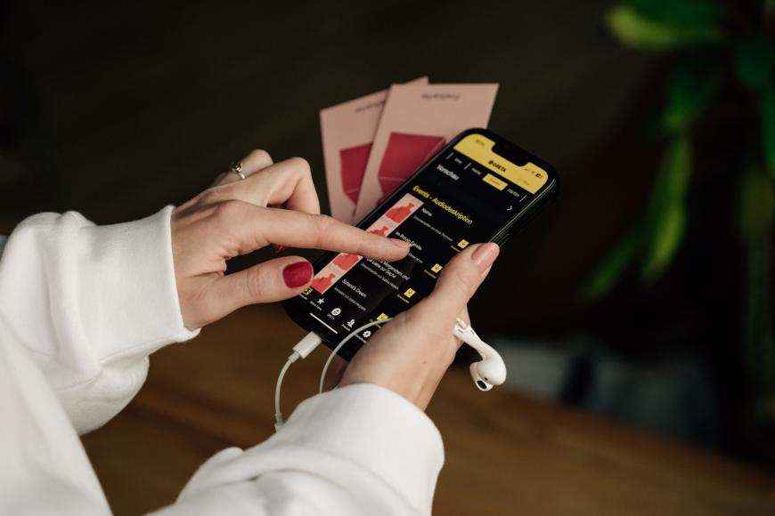 Eine Person hält ein Smartphone mit geöffneter GRETA-App in der Hand und tippt auf eine Audiodeskription.