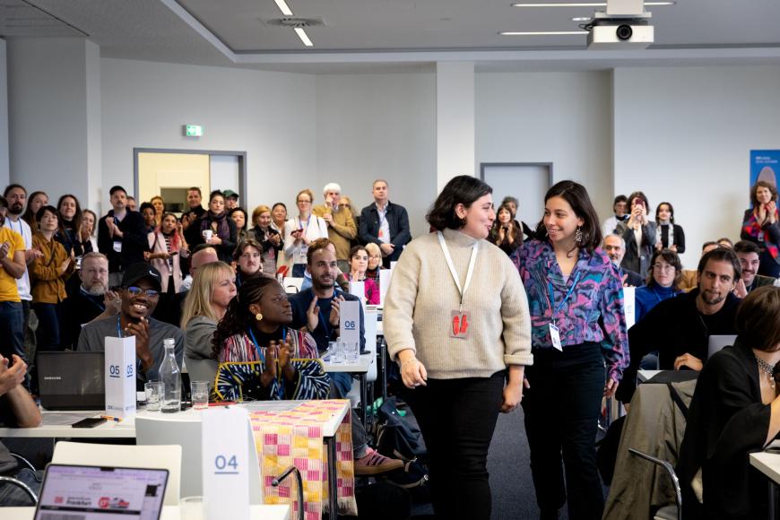 Two women are walking towards the camera in a crowded room, people are applauding