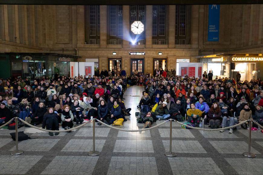 Die Osthalle des Hauptbahnhofs Leipzig ist voller Menschen, viele auf Stühlen, viele sitzen auf dem Boden, alle schauen gebannt zur Leinwand hinter der Kamera.