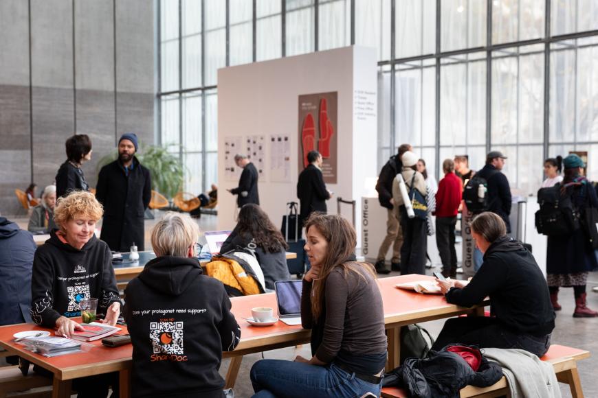Große, helle Halle mit langen Tischen, an denen viele Personen sitzen, Kaffee trinken und sich unterhalten. Im Hintergrund ist Gästecounter zu sehen und daneben eine Wand mit Festivalplakat und ausgedruckten Presseberichten.