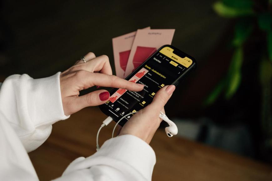 Nahaufnahme von Händen, die ein Handy halten, auf dem die Greta App für Audiodeskriptionen geöffnet ist